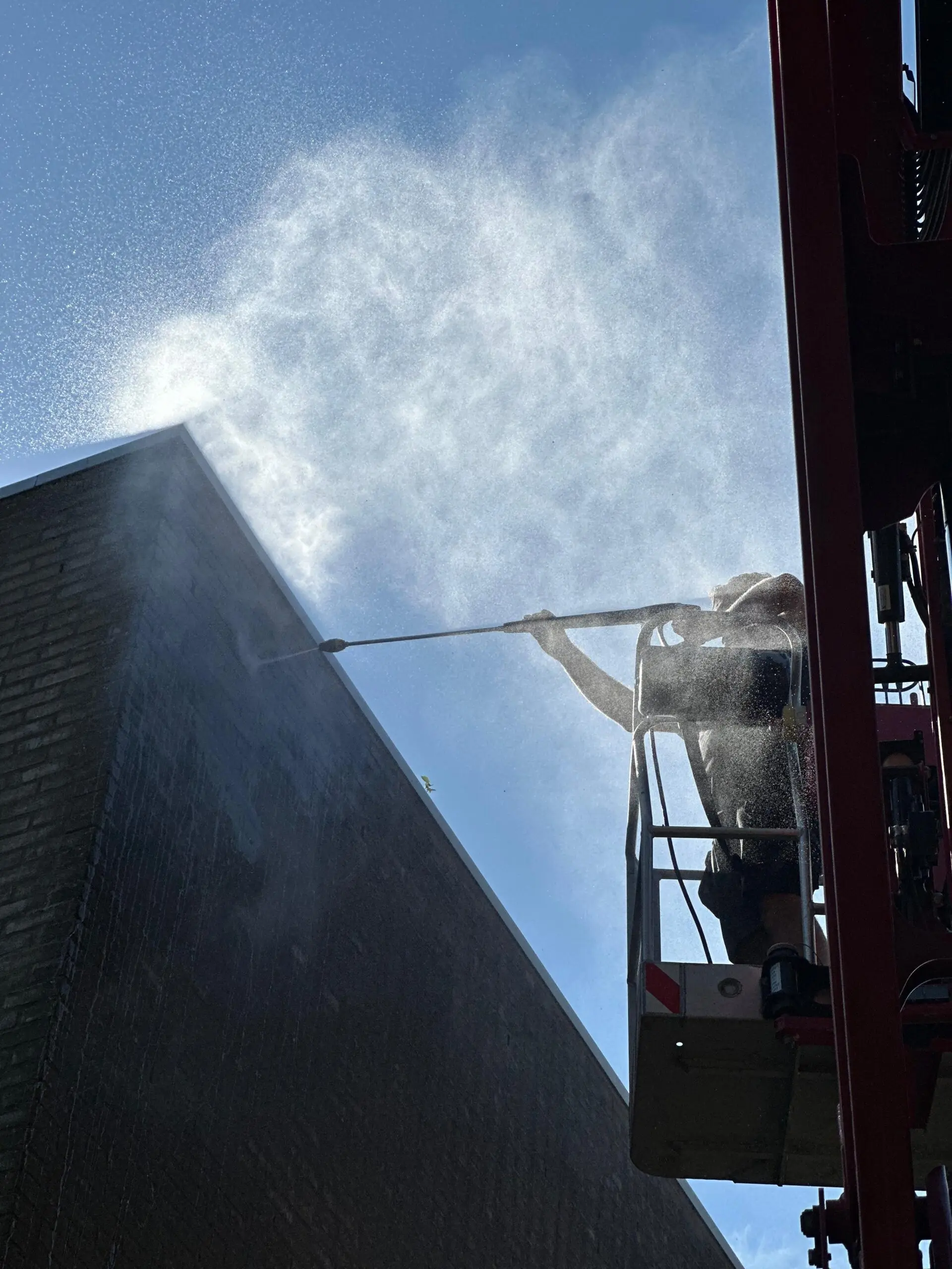 Een monteur van LEMA werkt vanuit een hoogwerker aan een gevel; de waternevel van de hogedrukspuit licht op tegen de blauwe lucht.